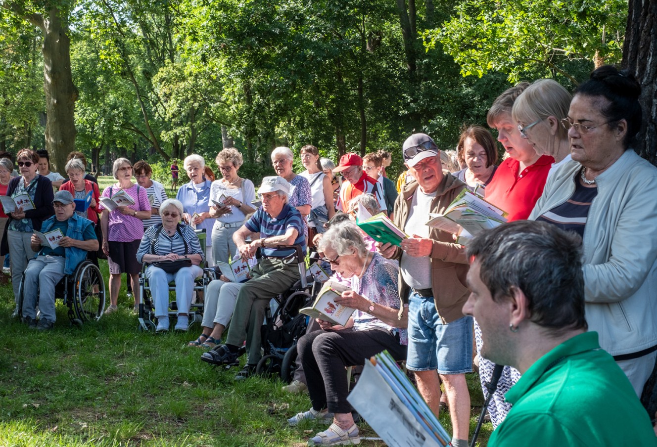 Eine Menschengruppe steht zusammen auf der Würfelwiese, einige Personen sitzen im Rollstuhl, viele halten Liederhefte in der Hand und singen